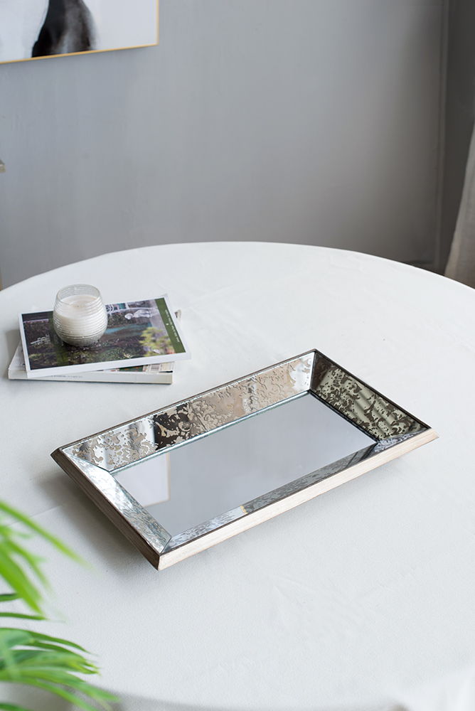 An Antique Rectangle Mirror With Floral Accents serves as a mirrored display tray on a round white table, next to two stacked books and a white candle in glass. A leafy green plant is seen in the foreground.