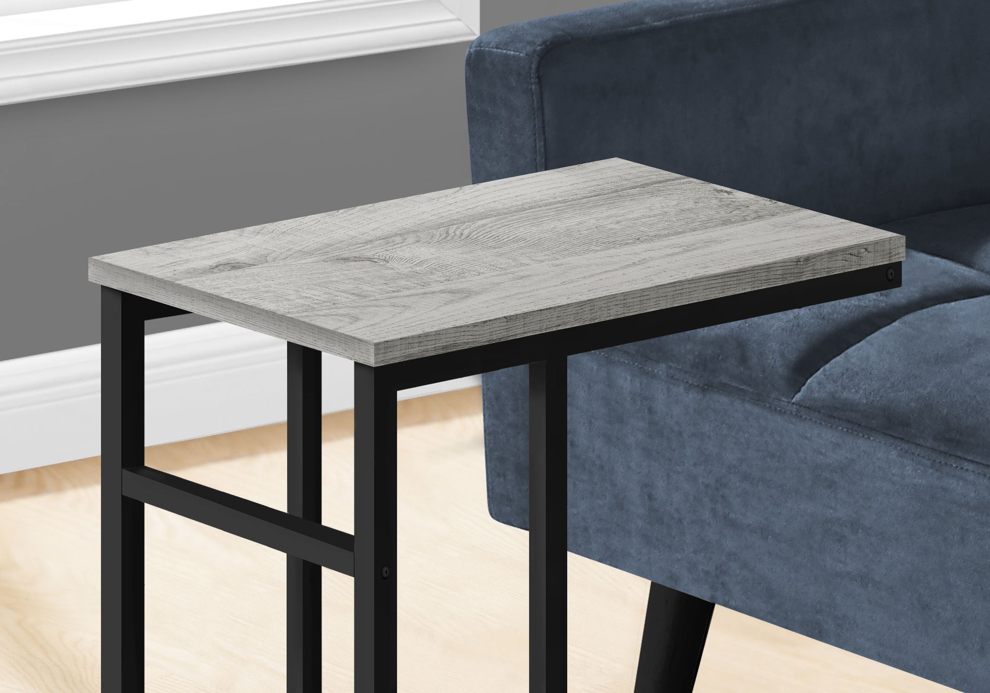 The Accent Table, C-Shaped with a light wood top and black metal legs sits beside a dark blue upholstered sofa in a room featuring gray walls and light wood flooring, adding a touch of contemporary style and versatile use.