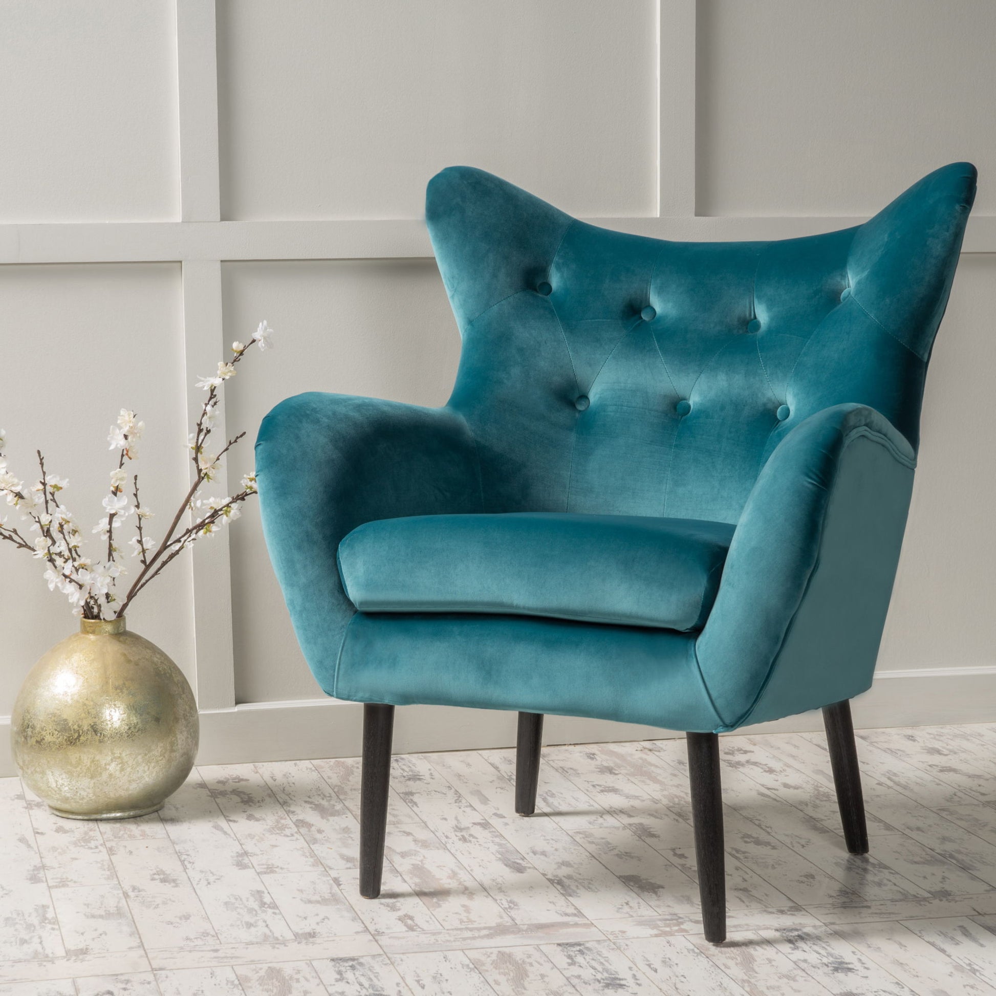 The Alyssa Mid Century Upholstered Arm Chair with wingback design in teal velvet and angled black legs sits on a light wood floor beside a gold vase of white flowers, set against a gray paneled wall.