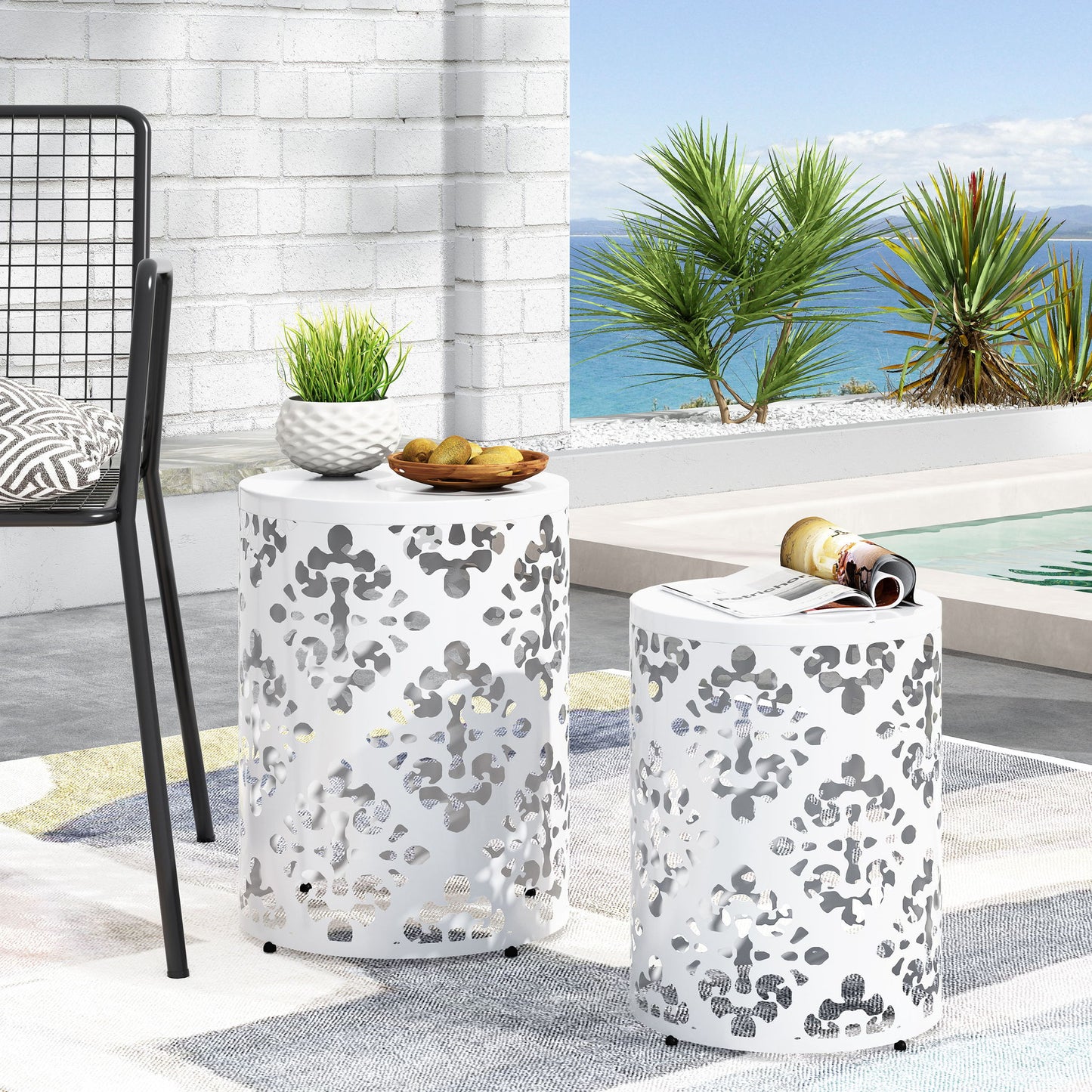 Two Outdoor Metal Modern Design End Tables with cut-out patterns sit beside a pool—one holds a plant and snacks, the other a magazine. Palm trees and an ocean view are visible in the background.
