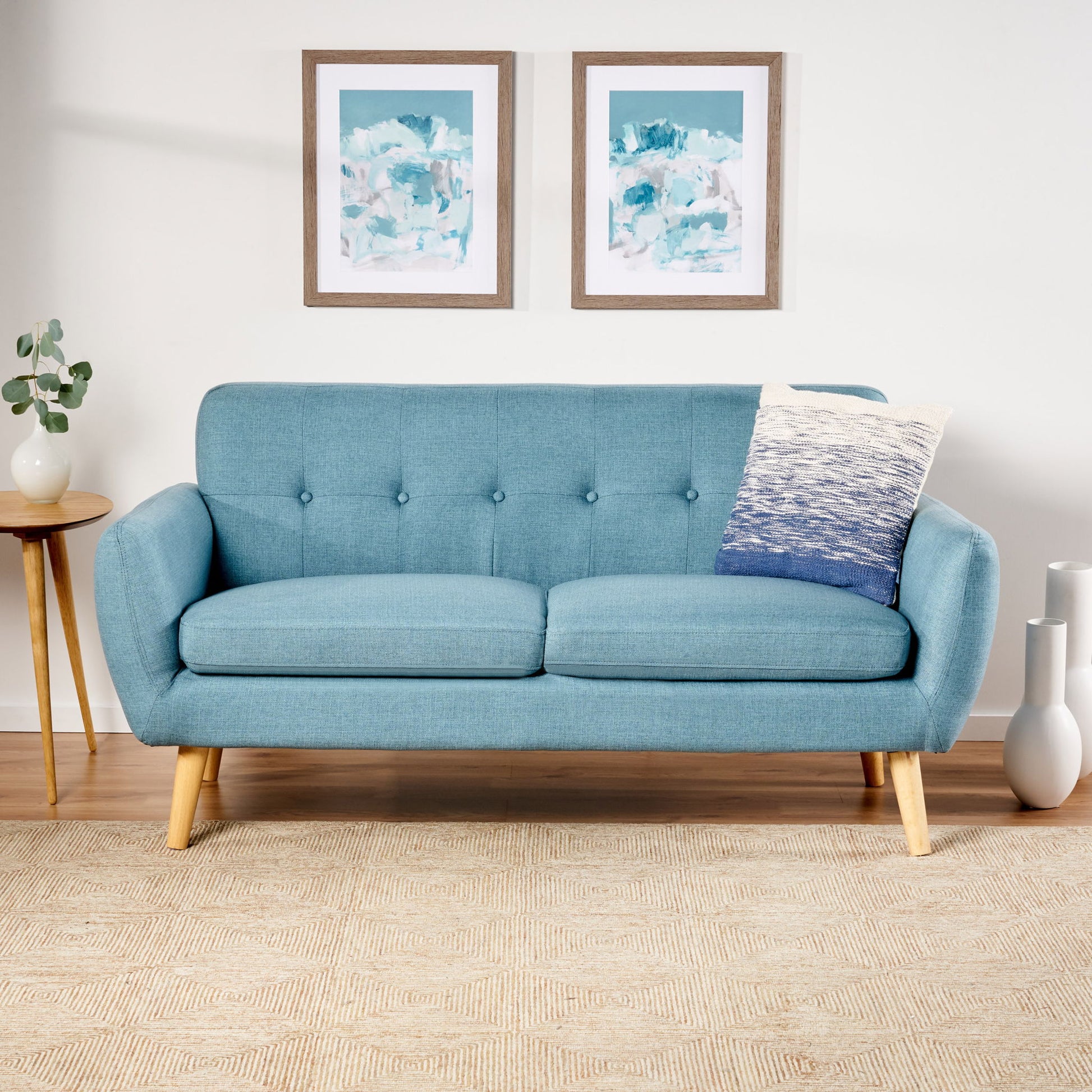 A Modern Loveseat Sofa With Button Tufted Backrest in light blue stands in a living room with wooden legs, paired with a gradient pillow. A round side table holds a plant, while two abstract art pieces hang above on the wall.