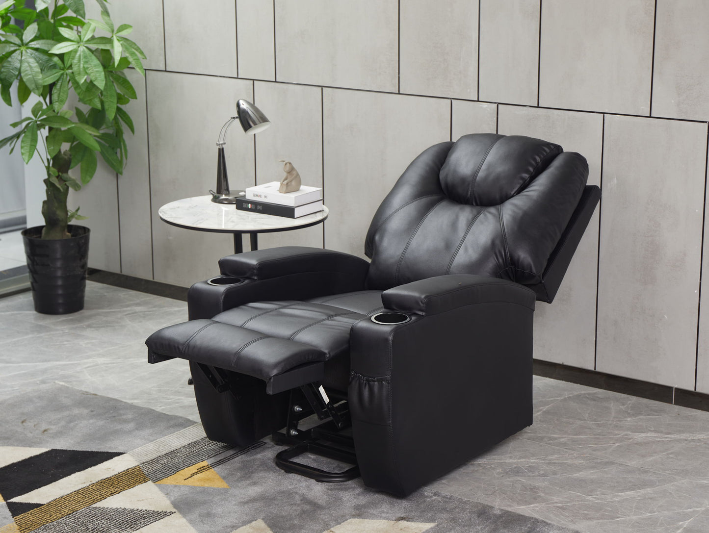 A Modern Recliner Chair With Cup Holders and Remote Control sits beside a round marble side table with a lamp, book, and small sculpture, set against a tiled wall near a potted plant.