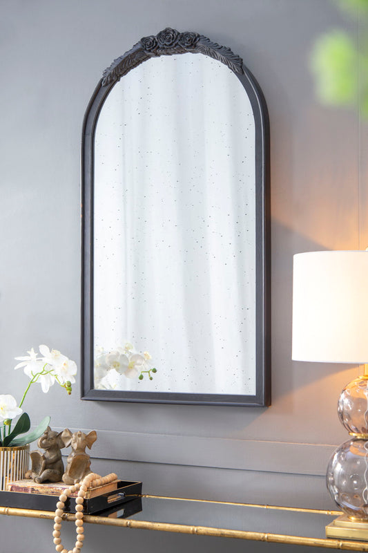 A Hand Carved Rose Antique Mirror Frame, Wood Arch Mirror Wall Decor for Living Room, hangs on a gray wall above a console table displaying a lamp, books, beaded garland, elephant sculpture, and white orchids in a vase.