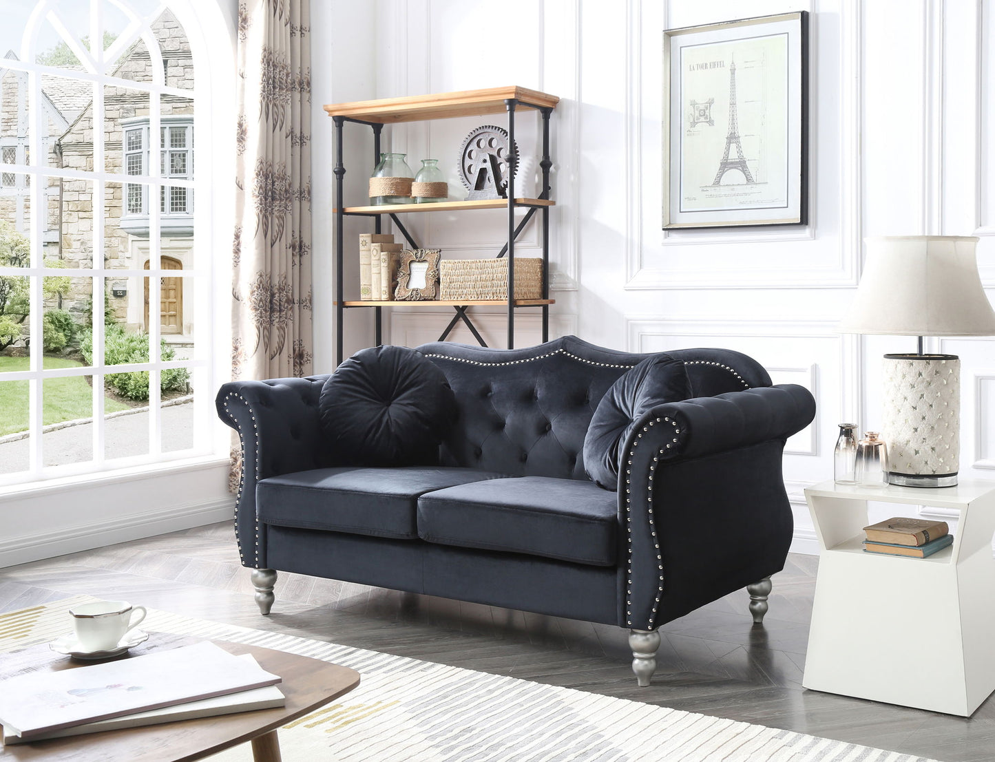 A cozy living room featuring a Stylish Flared Arm Loveseat in dark blue, decorative pillows, a white side table with a lamp, books and decor on shelves, and a large window allowing natural light.