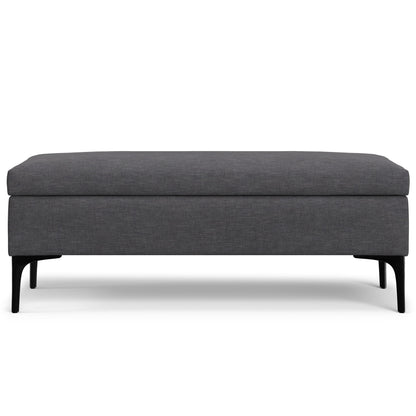 The Rebecca Multifunctional Coffee Table Storage Ottoman features a rectangular, dark gray upholstered design with four slender black legs, shown from the front against a white background.