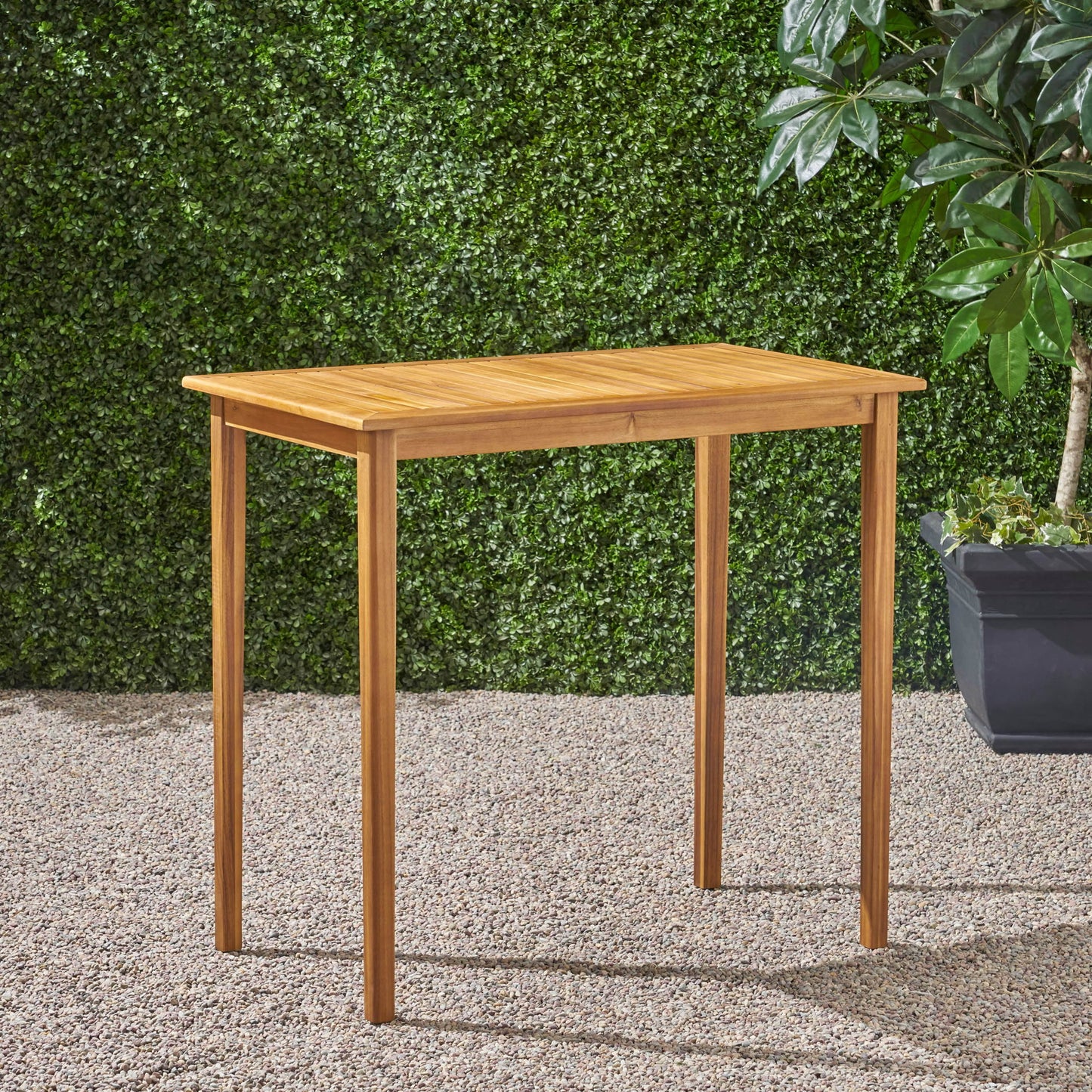 The Outdoor Minimalist Acacia Wood Rectangle Bar Table with four straight legs stands on gravel, set against a green leafy hedge and accompanied by a potted plant in the background.