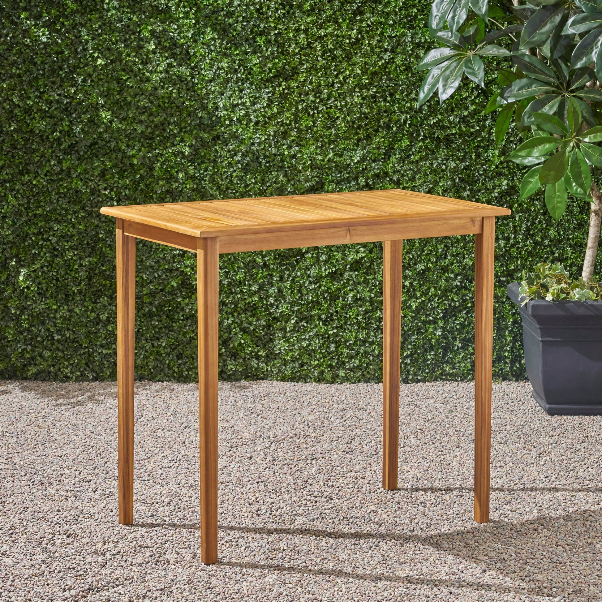 The Outdoor Minimalist Acacia Wood Rectangle Bar Table with four straight legs stands on gravel, set against a green leafy hedge and accompanied by a potted plant in the background.