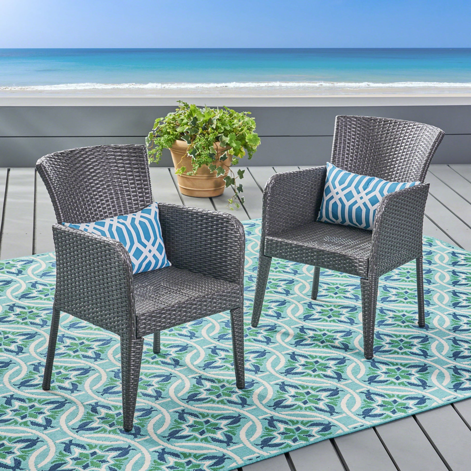 Two Anaya Outdoor Wicker Dining Armchairs with geometric blue cushions sit on a patterned outdoor rug on a deck overlooking the ocean, separated by a potted plant, with clear skies and calm sea in the background.