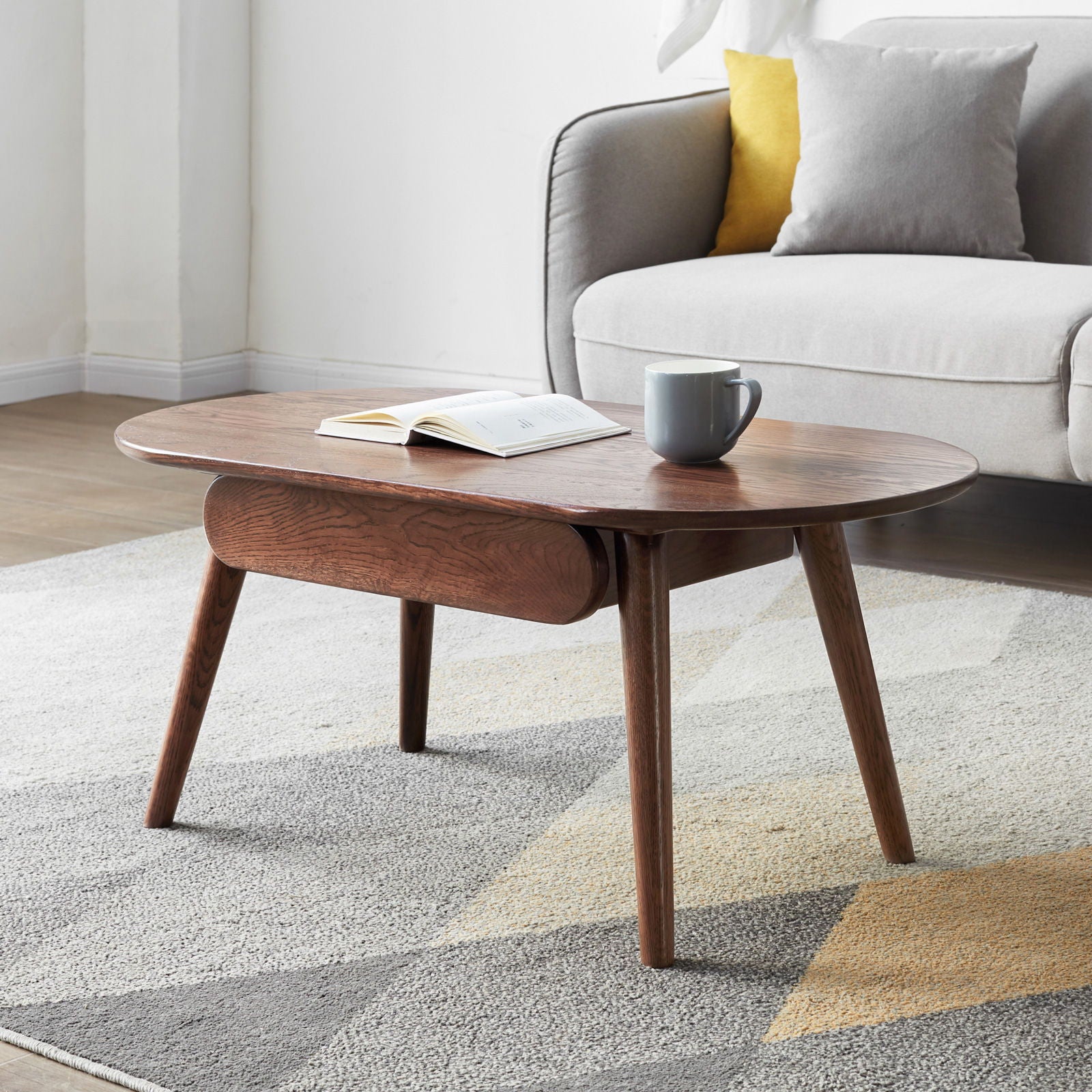 The Capsule Centre Low Table With Drawers Coffee Table, topped with an open book and gray mug, stands on a patterned rug in front of a light gray sofa adorned with gray and yellow cushions in a modern living room.