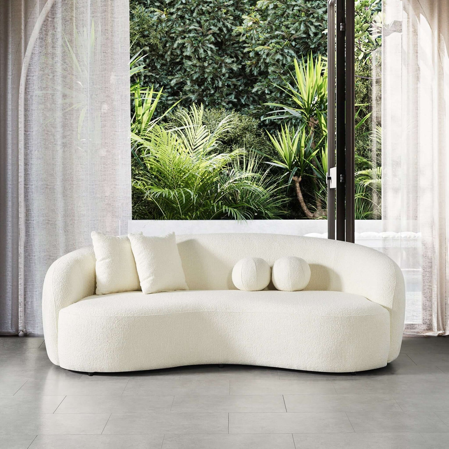 The Drake - Japandi Style Curvy Boucle Sofa in modern white, featuring three cushions, sits on a gray tile floor before large windows with sheer curtains, overlooking lush greenery.