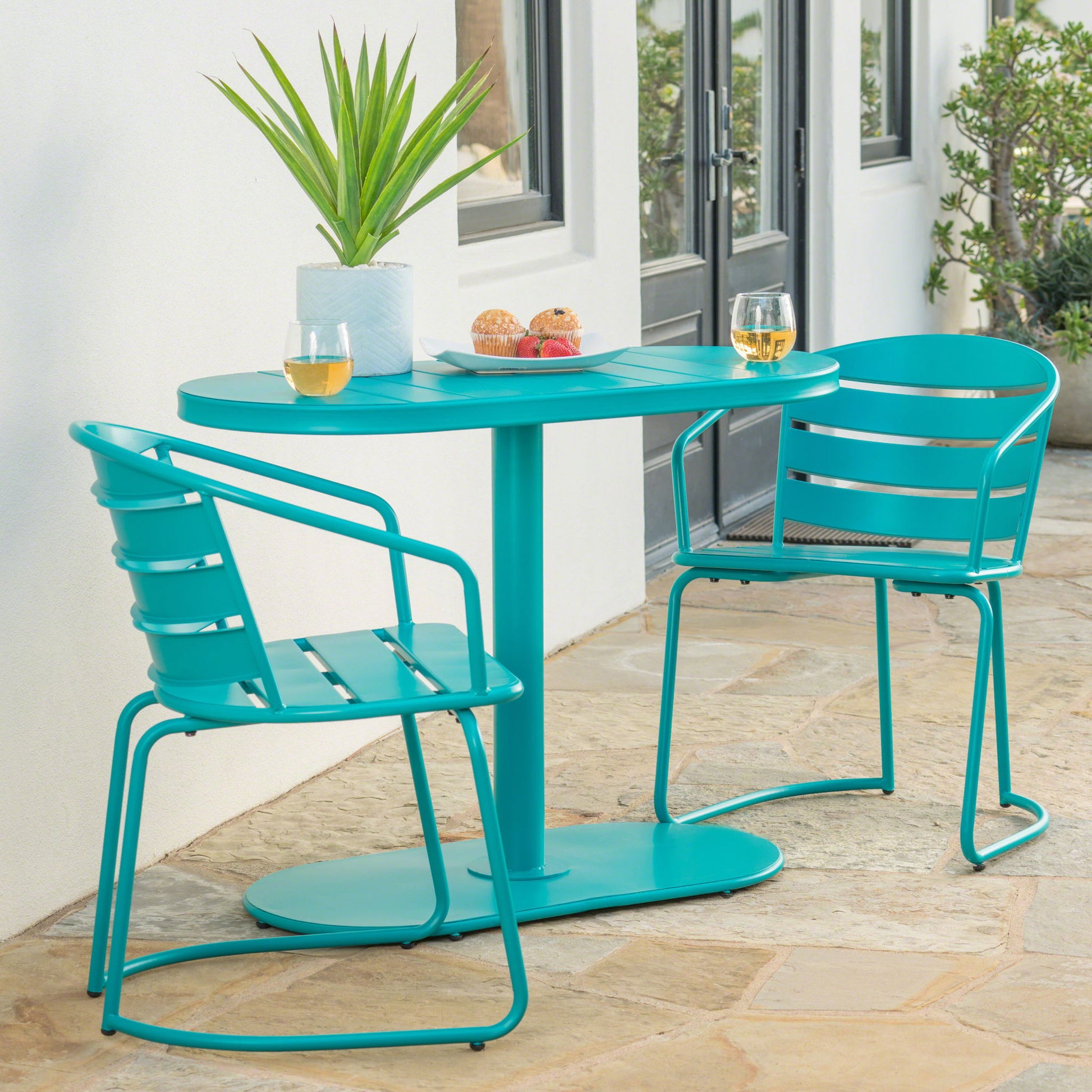 A 3 Piece Elegant Outdoor Bistro Set with 2 chairs and an oval table is displayed on a stone patio beside a white building, the table set with two glasses of white wine, pastries, and a potted plant.