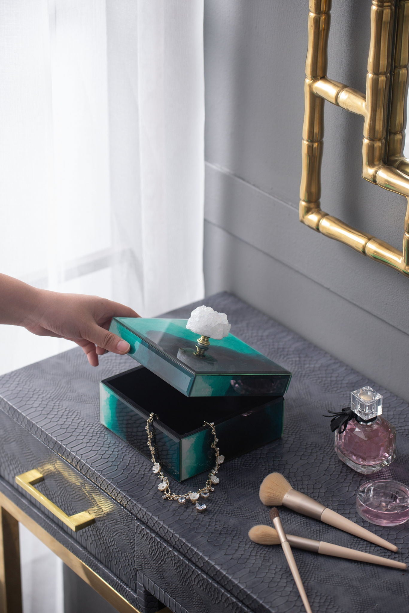 A hand opens the Bethany - Aqua Jewelry Box, a stackable decorative storage box with a crystal knob, on a textured gray table revealing a necklace. Nearby are perfume, makeup brushes, and a gold-framed mirror by the gray wall.