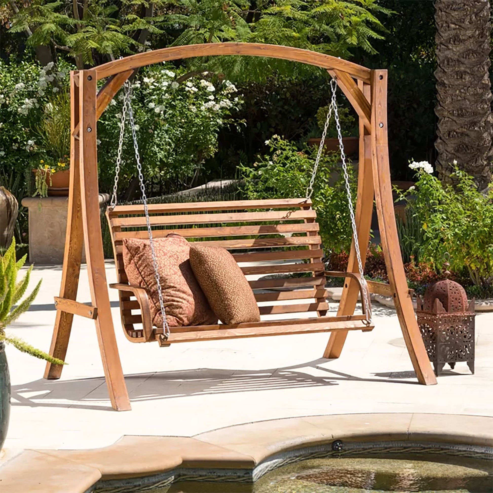 The San Juan - Outdoor Swinging Loveseat With Support hangs on a sunny patio, featuring three decorative pillows. It’s set among lush greenery, stone tiles, and overlooks a small pool in the foreground.