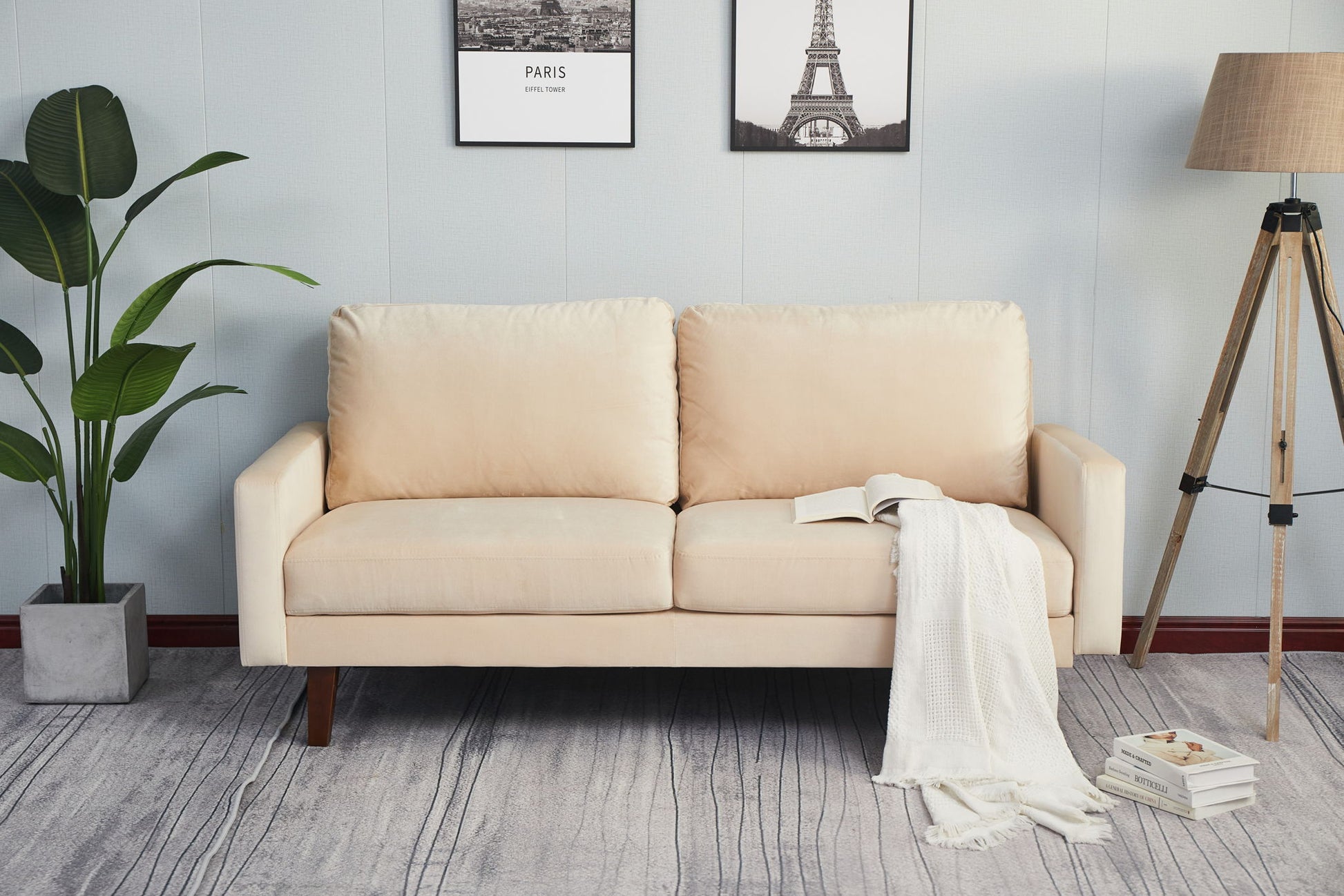 A Comfortable Sofa With Durable Construction, in beige with cushions, is featured in a living room styled with a white throw, books on the floor, a potted plant, tripod floor lamp, and Paris-themed framed art.