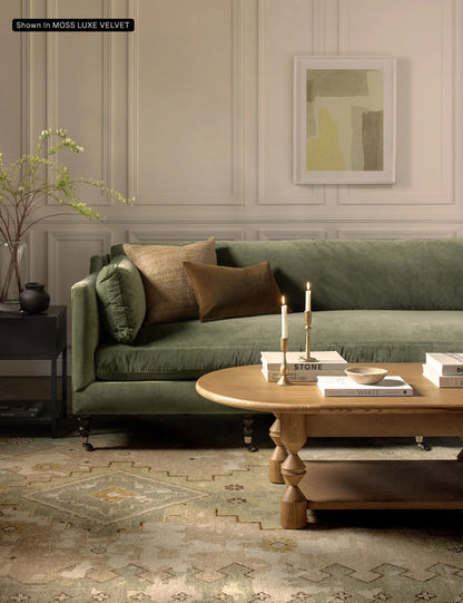 A cozy living room featuring the Fabienne Sofa in green velvet, adorned with brown and beige pillows, complemented by a wooden coffee table. Two lit candles and books rest on the table, while a potted plant and an abstract painting add decorative touches to the space.