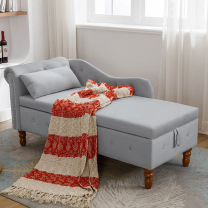 The Chaise Lounge Indoor, a modern velvet lounge chair with storage, rolled arms, nailhead trim, and pillow, sits by a window. A red and beige striped blanket and matching pillow are atop it; light curtains and a patterned rug complete the cozy scene.
