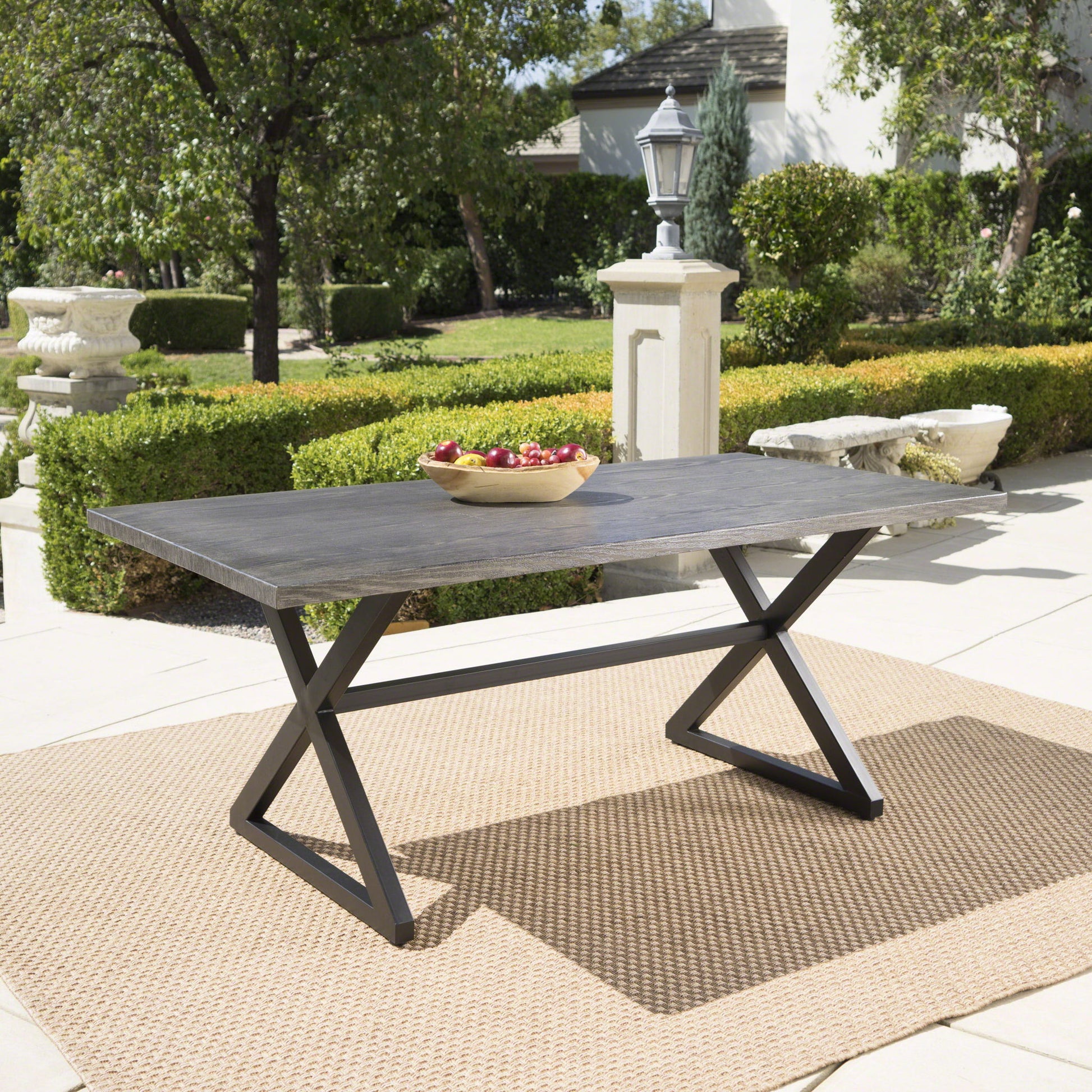 The Rolando Outdoor Aluminum Dining Table with Steel Frame sits on a tan woven rug in a landscaped garden. A wooden bowl of red apples rests on its dark rectangular top, surrounded by lush greenery and trees.