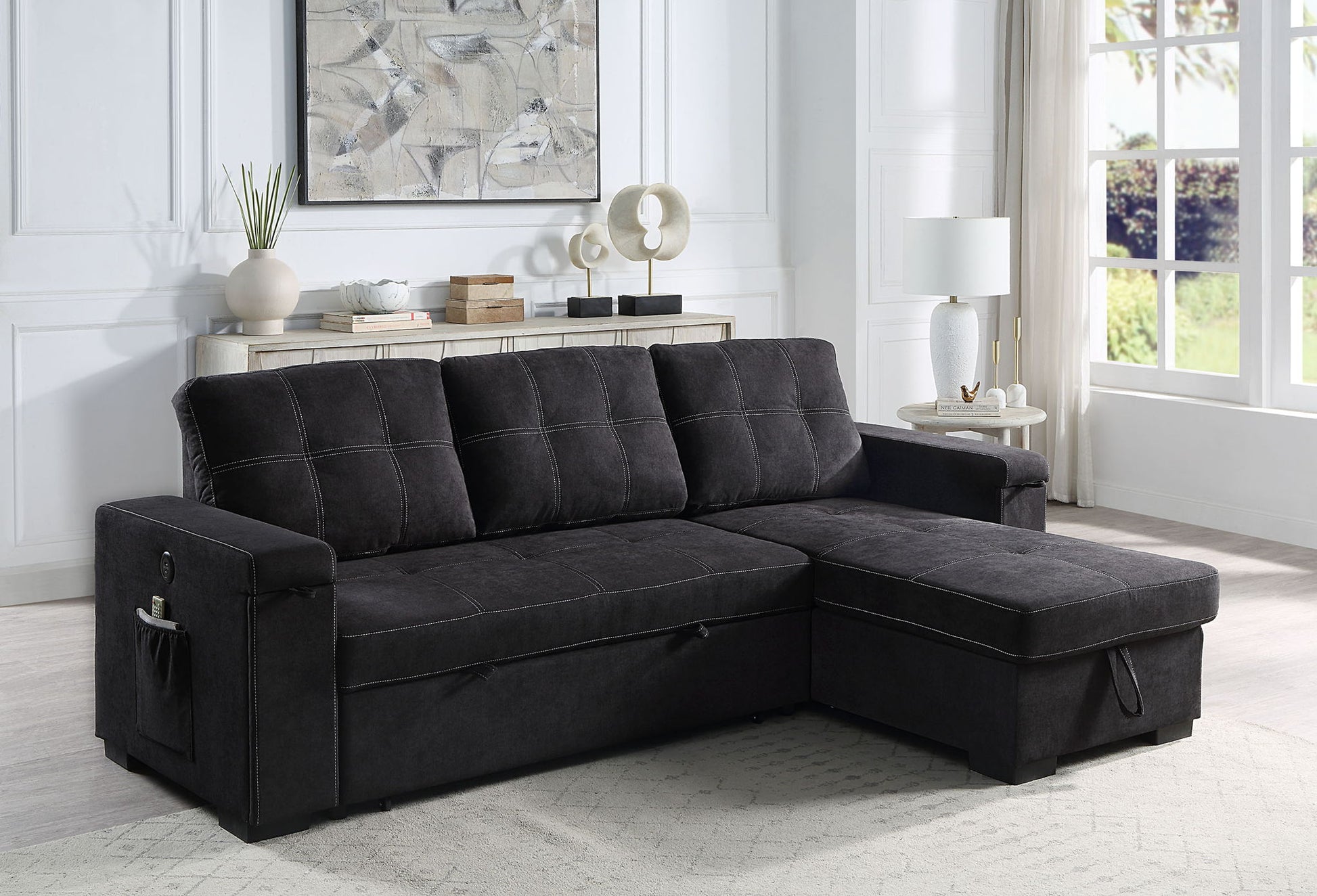 A modern living room with the Toby woven fabric reversible sleeper sectional sofa featuring storage, cup holder, charging ports, and pockets, paired with a light area rug, floor lamp, and console table in front of a large sunlit window.