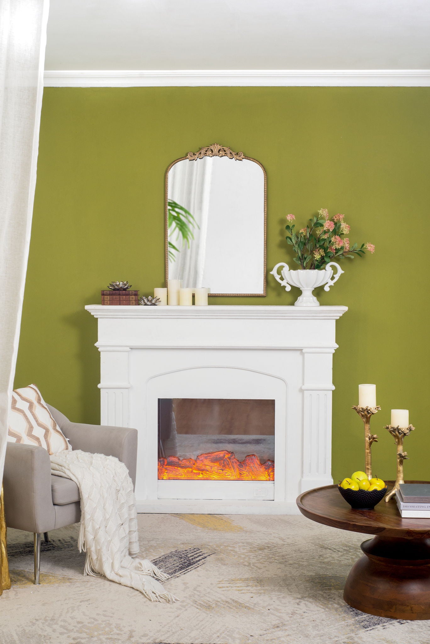 A cozy living room with an olive green wall, a white fireplace, and a Baroque-inspired Arch Mirror above the mantel. Candles and a flower vase sit on the mantel, while a gray chair with blanket and round table with candles and lemons complete the setup.