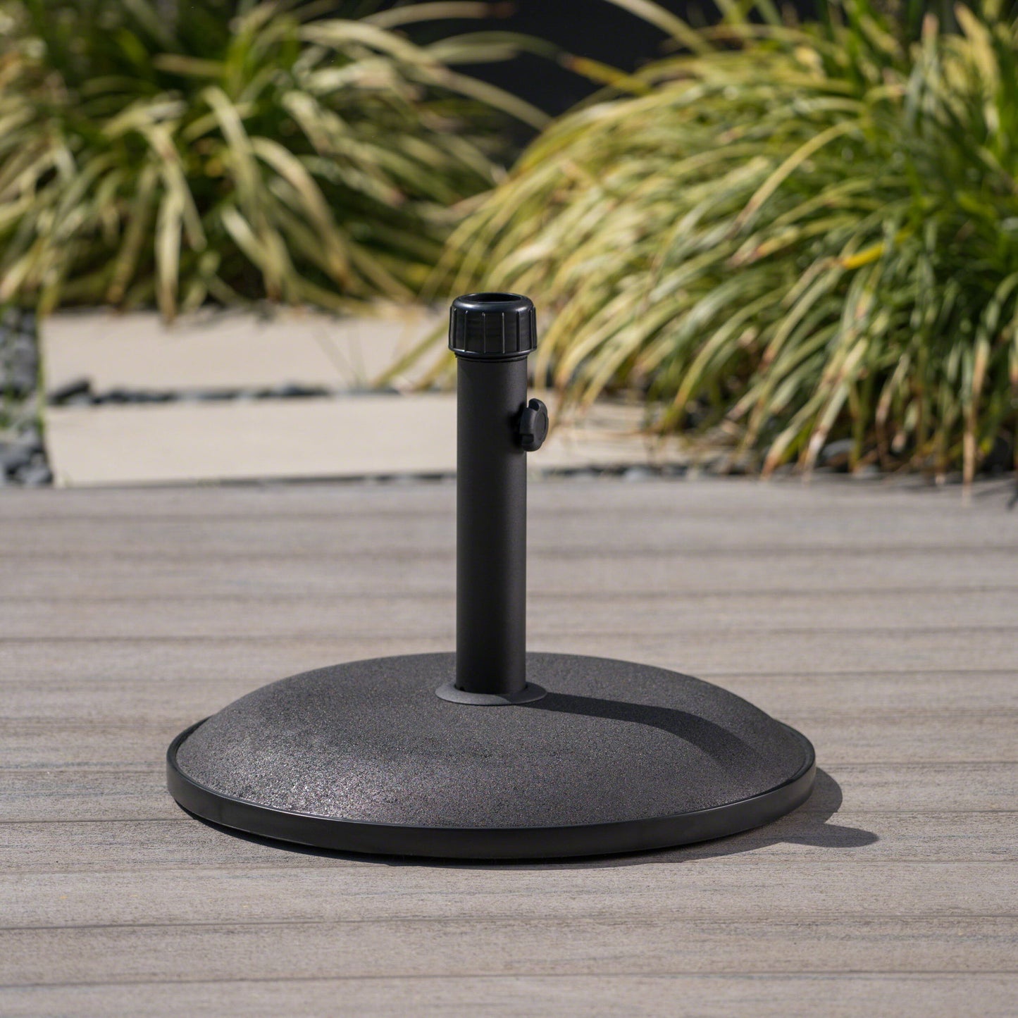 The Stylish Concrete Umbrella Base With Iron Support Pole - Black sits on a wooden deck, its vertical pole and tightening knob visible, with green ornamental grasses softly blurred in the background.