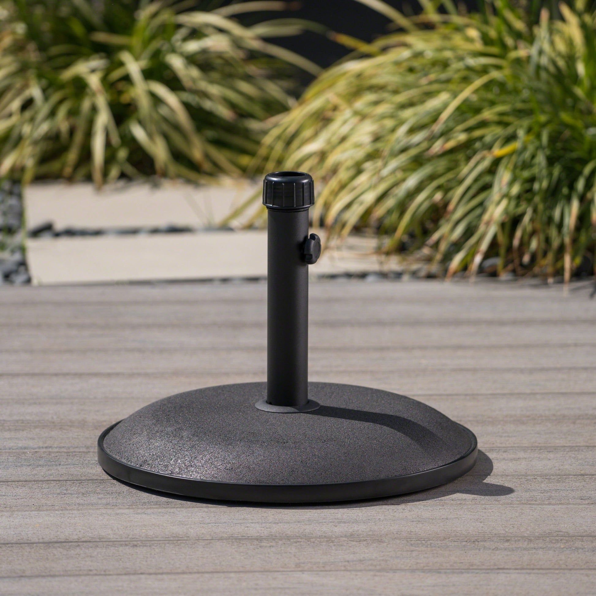 The Stylish Concrete Umbrella Base With Iron Support Pole - Black sits on a wooden deck, its vertical pole and tightening knob visible, with green ornamental grasses softly blurred in the background.