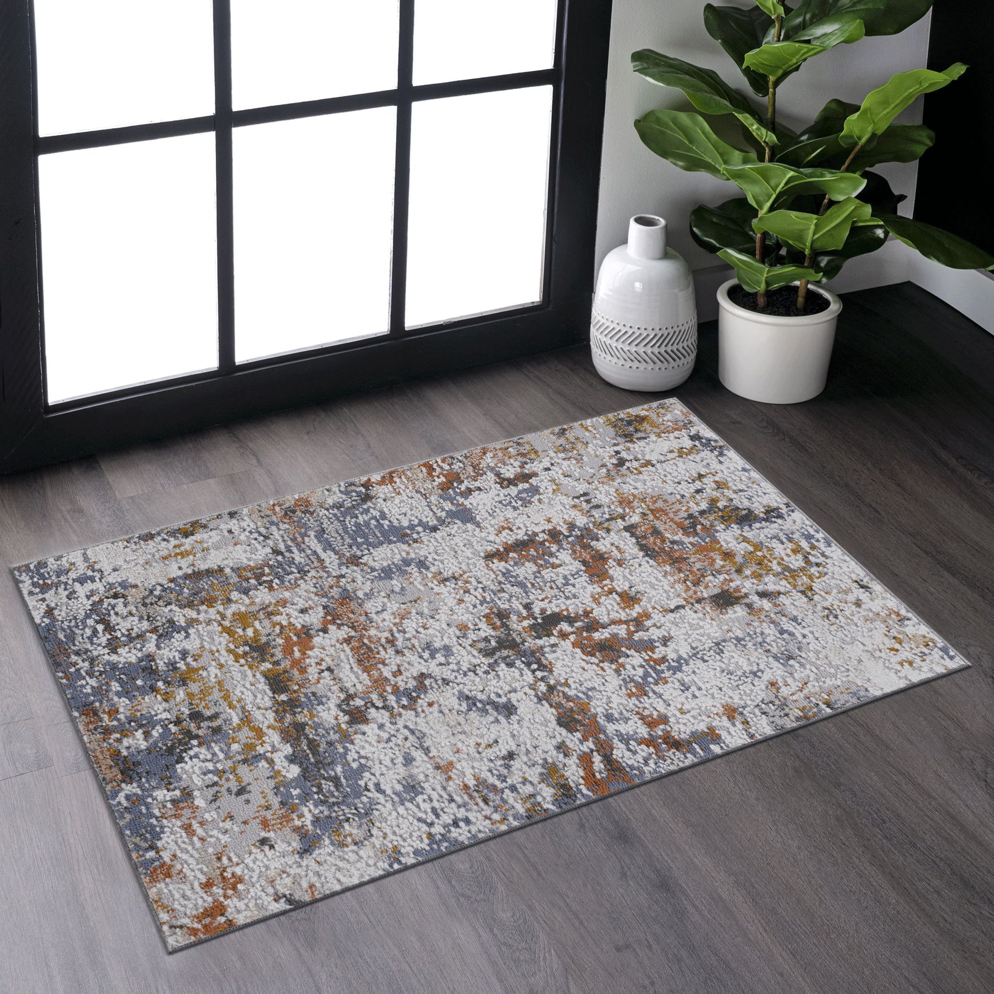 The Payas - Abstract Area Rug, featuring gray, white, and orange abstract patterns, is placed on a dark wood floor by a large window, with a potted plant and white decorative vase in the corner.