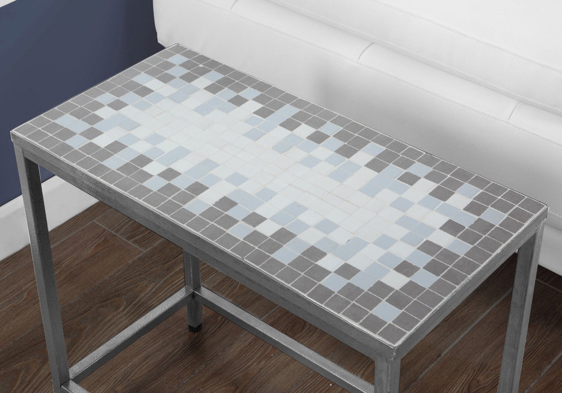 The Perfect Accent Table, Side Transitional features a rectangular metal base and a mosaic tile top in gray, white, and light blue with a pixelated design. It’s shown on a dark wood floor beside a white couch and navy blue wall.