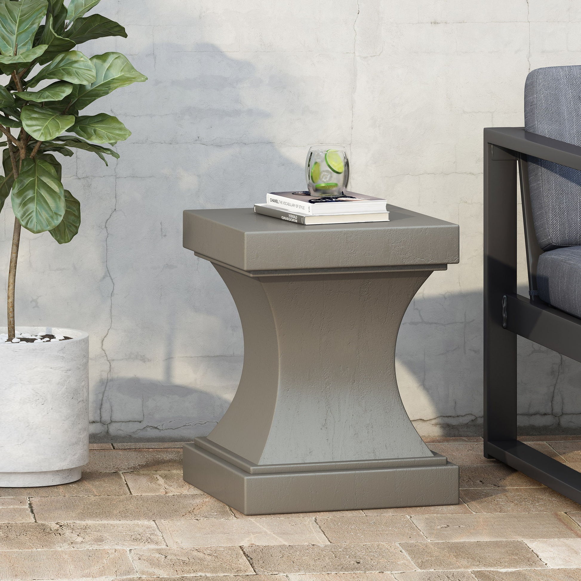 The Athena - Modern Side Table With Pedestal Design, in gray, sits on a brick patio next to a potted plant and dark outdoor sofa. On top are a glass of water with lime and two books, set against a concrete wall.