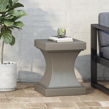 The Athena - Modern Side Table With Pedestal Design, in gray, sits on a brick patio next to a potted plant and dark outdoor sofa. On top are a glass of water with lime and two books, set against a concrete wall.