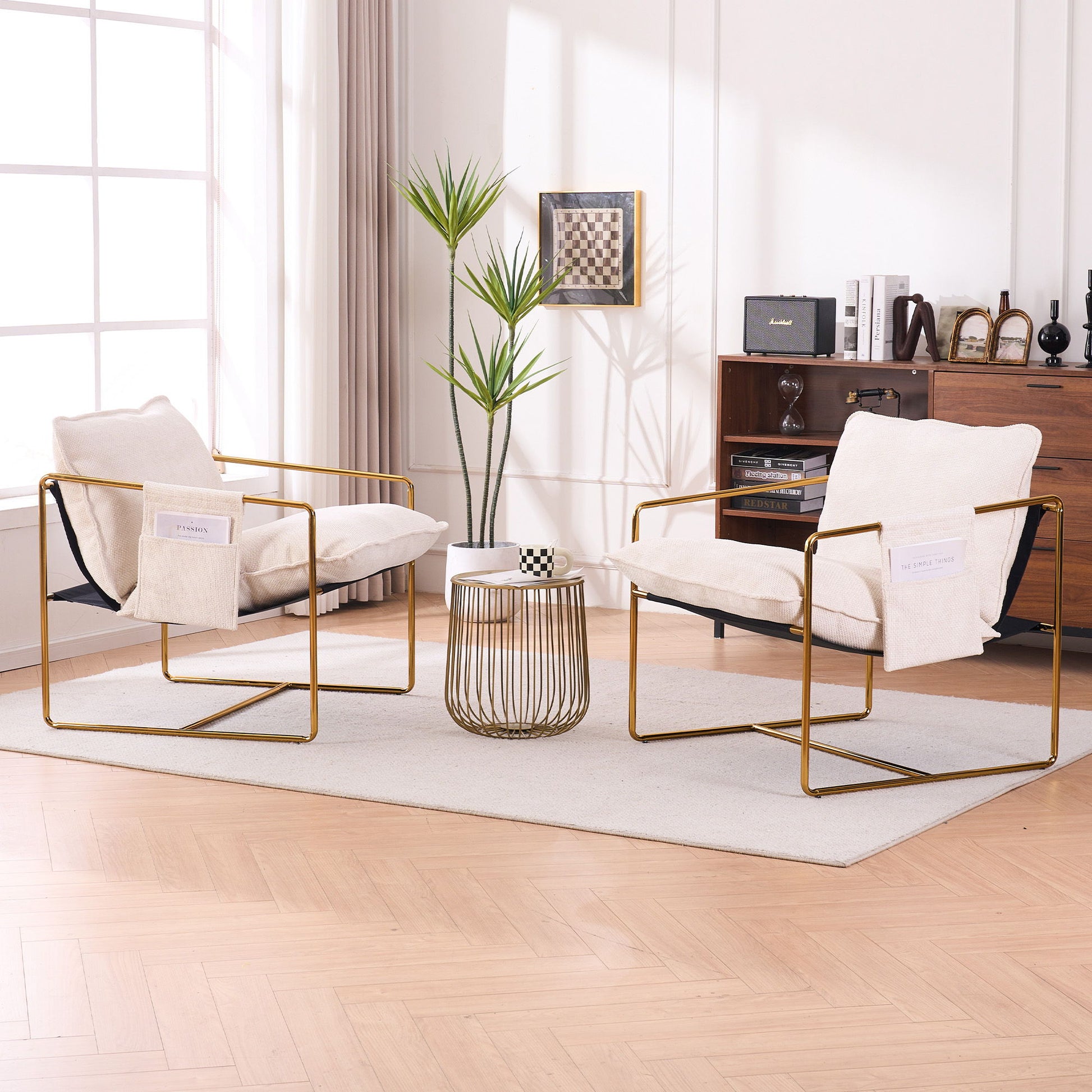Two Upholstered Hanging Armchairs with gold-plated metal frames, crushed foam cushions, and skin-friendly woven fabric face each other in a bright living room. A round side table sits between them, with decor and sunlight streaming through large windows.