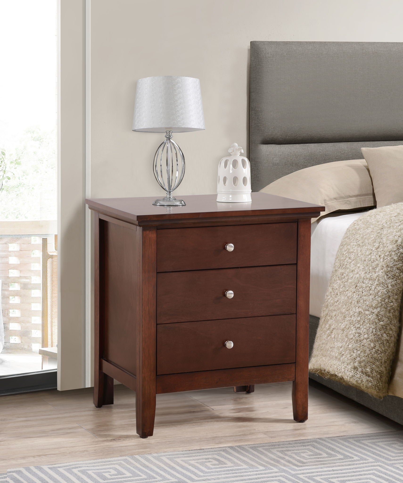 The Hammond - 3 Drawer Nightstand with silver knobs stands by the bed. On top sit a modern silver lamp with a white shade and a small white decor piece, complementing the room’s neutral tones and cozy feel.