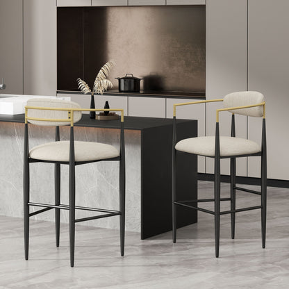 A set of two Counter Height Upholstered Barstools with black frames, gold armrests, and beige cushions sits by a marble kitchen island in a minimalist space with neutral cabinets and a decorative vase.