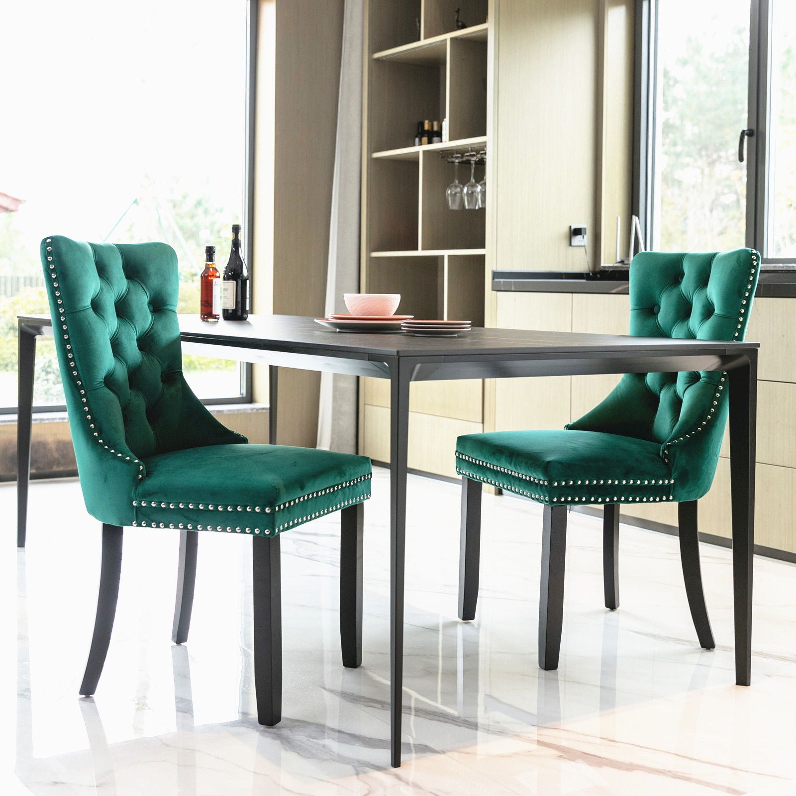 Two Nikki tufted velvet dining chairs with nailhead trim stand at a black rectangular table in a bright, modern kitchen with large windows and marble floors. Wine bottles and glasses decorate the table and shelves.