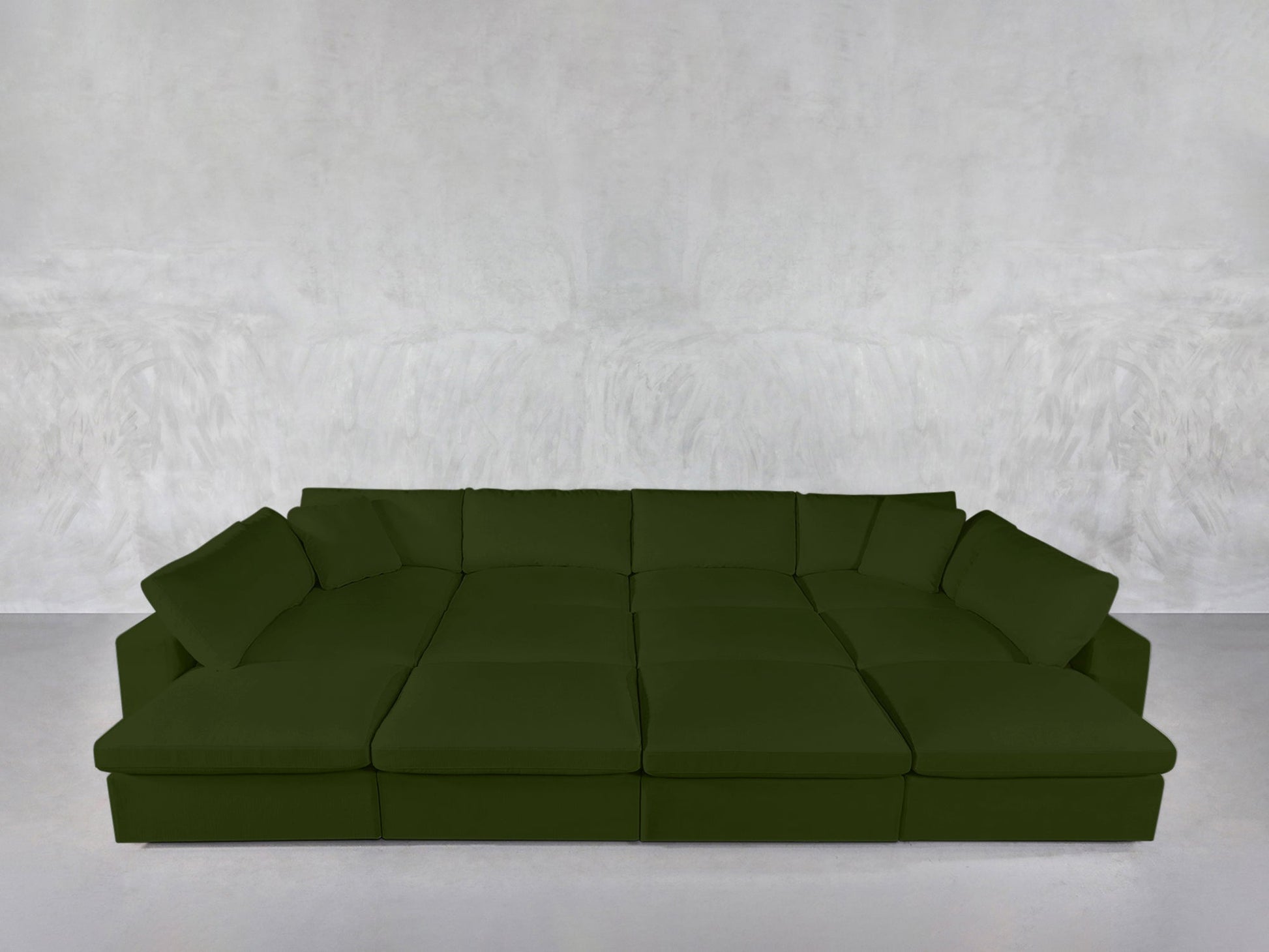 The 12-Seat Deep Modular Daybed, in deep green with multiple seat cushions and back pillows, stands against a light gray textured wall in a minimalistic room with a pale floor.