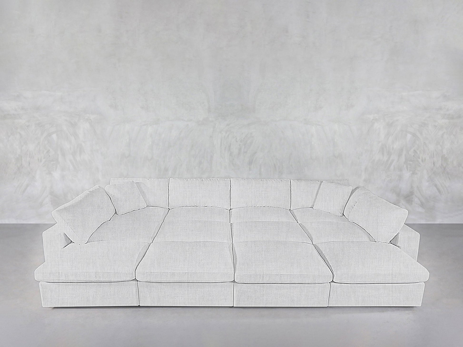A spacious, modern 12-Seat Deep Modular Daybed with plush cushions and matching pillows is arranged against a plain, light gray textured wall and floor.