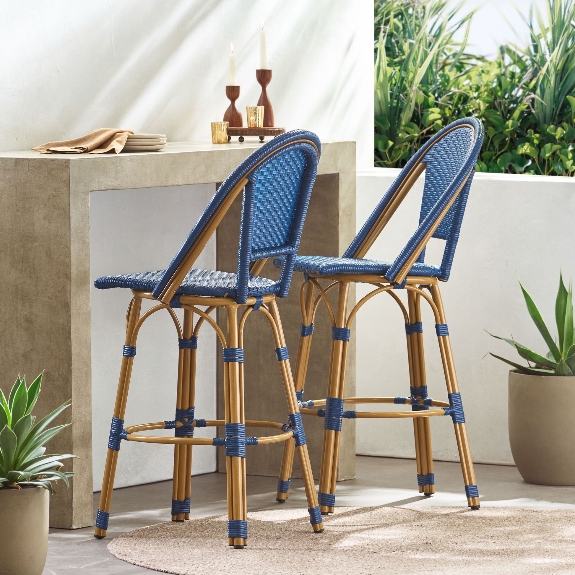Two Outdoor Rattan And Aluminum French Barstools (Set of 2) sit at a modern concrete bar table with candlesticks, plates, and napkins. The scene features green plants, natural light, and a beige round rug.
