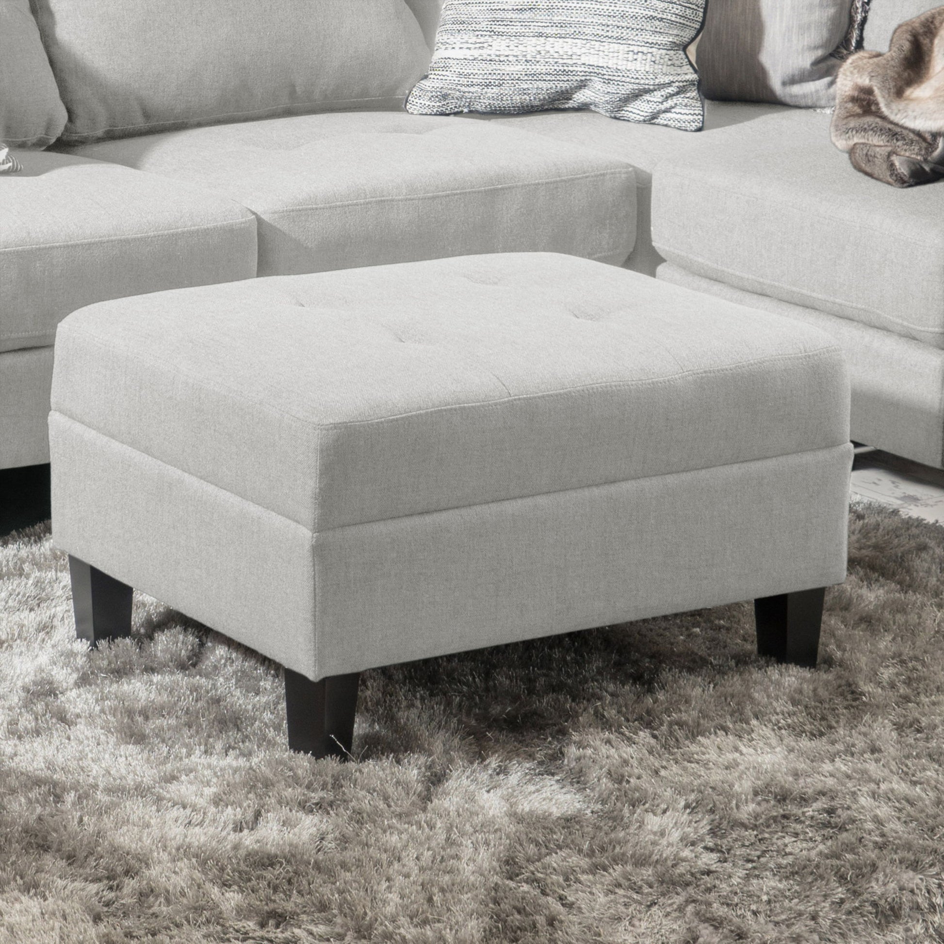 A Classic Ottoman With Birch Wood Legs And Fabric Upholstery sits on a textured gray rug in front of a matching sectional sofa with pillows and a blanket.