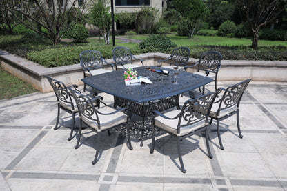 The Square 8 Person 64" Long Aluminum Dining Set With Cushions, in black, features eight chairs around a square table on a tiled patio, with a book and flowers on the tabletop and green shrubs and trees in the background.