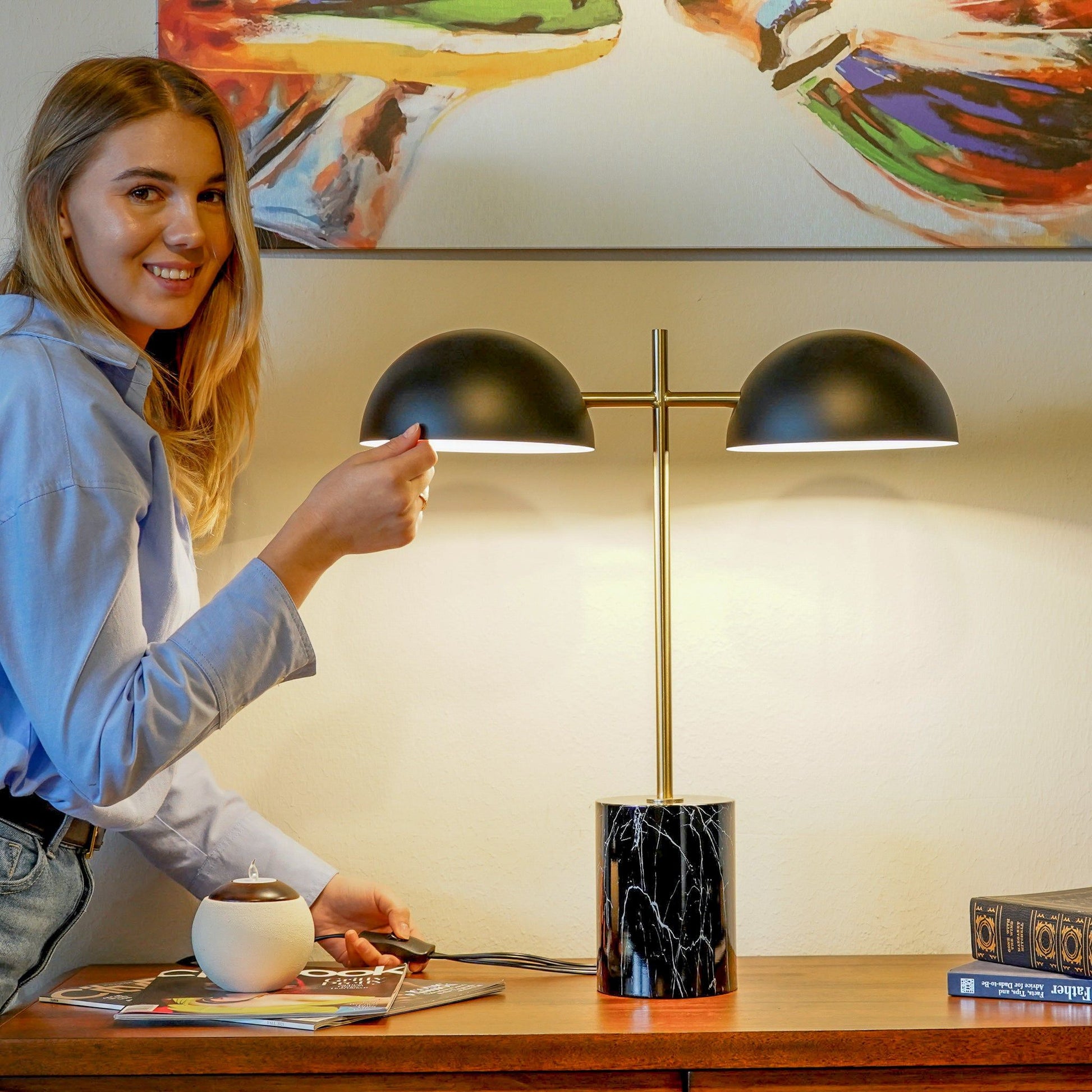 A woman in a blue shirt smiles as she turns on the Nova - Hydro Table Lamp With On / Off Switch Double Lamp in Black, Gold, and Marble atop a wooden table with a mug, book, and decor beneath an abstract painting.