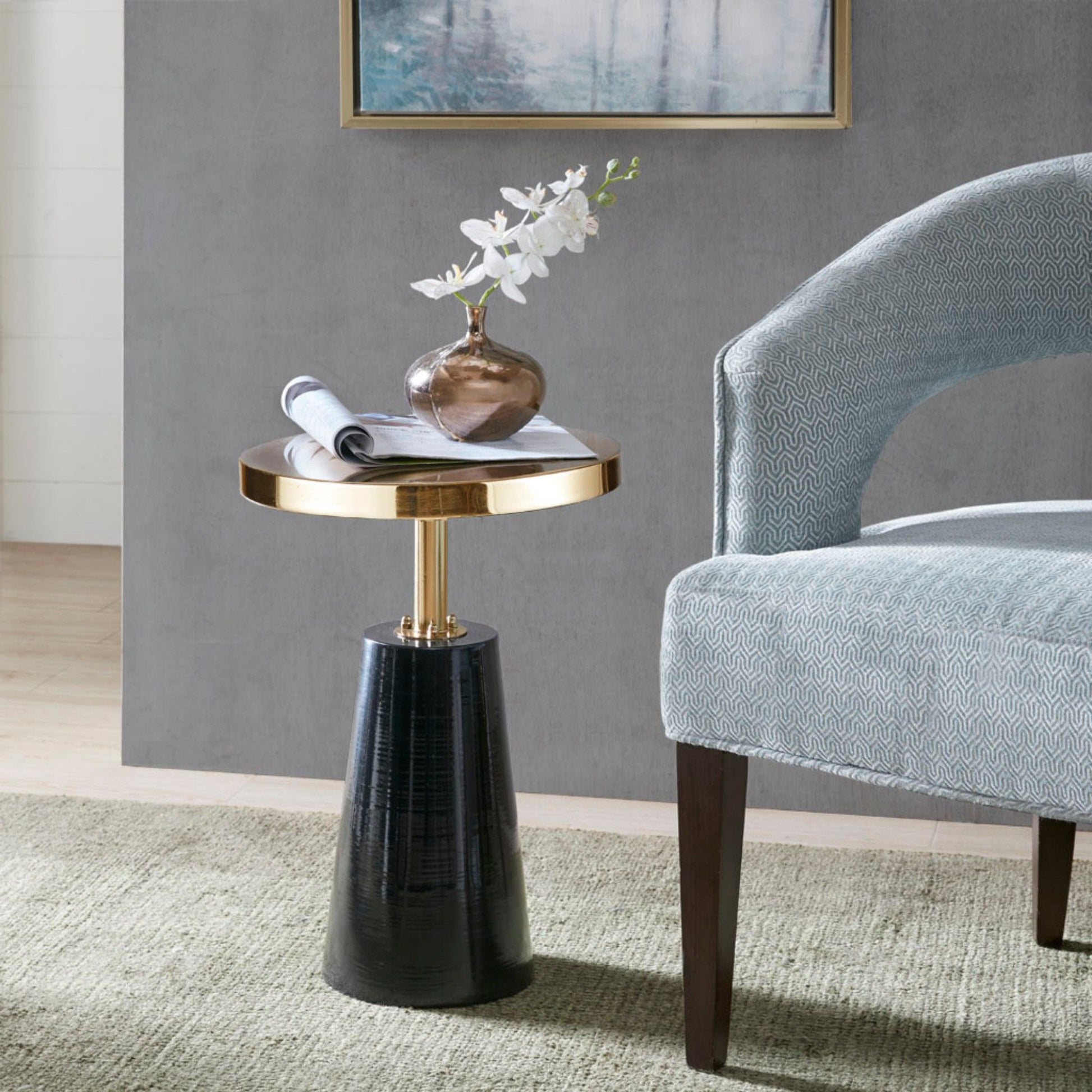 The Accent Table Elegant Design Metal Round - Black / Gold holds a vase with white flowers and a rolled magazine, placed beside a light blue upholstered chair on a green rug, set against a gray wall with framed art.
