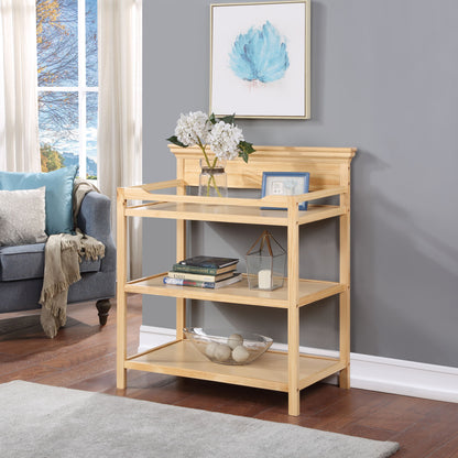 A Universal - Changing Table in light wood with three tiers stands against a gray wall, styled with books, a vase of white flowers, a photo frame, and a glass bowl beside a blue-gray sofa in a cozy living room.
