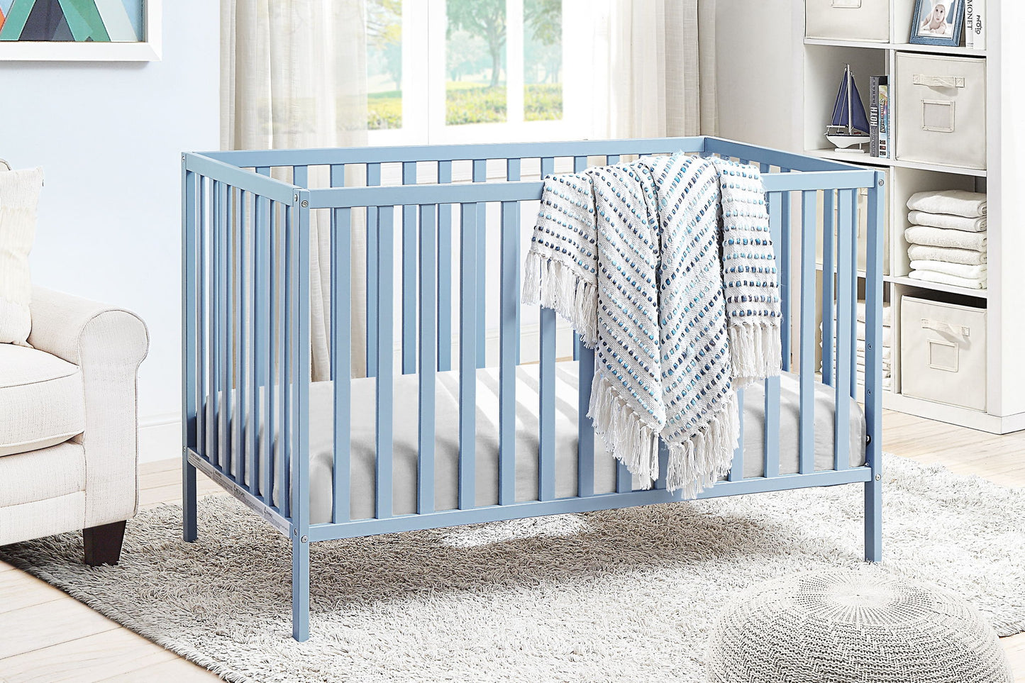 The Palmer - 3-in-1 Convertible Island Crib in light blue, featuring a white and blue patterned blanket, is set in a bright nursery with a beige armchair, shelving, and a large window in the background.