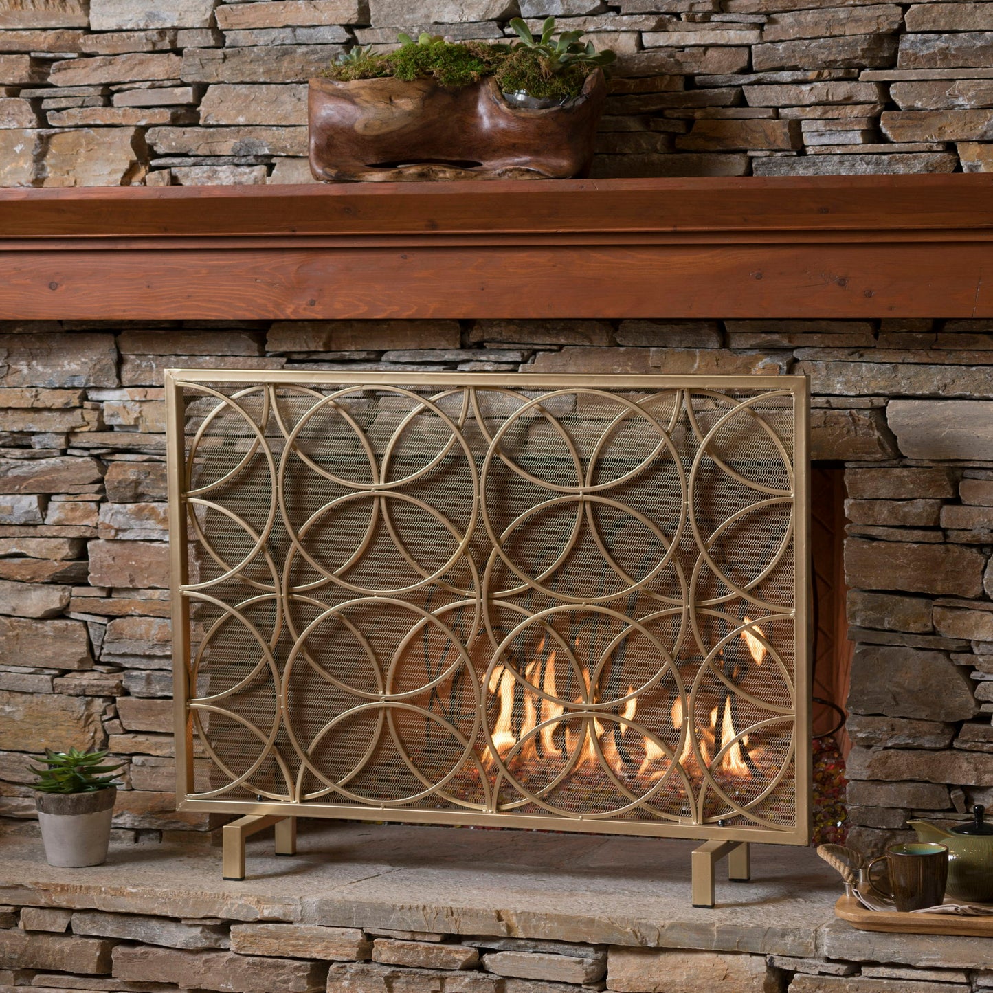 The Thayer Single Panel Iron Fireplace Screen with geometric design stands before a stone wall and wooden mantel, with glowing flames behind it. Potted plants and a teacup rest on the mantel and hearth.