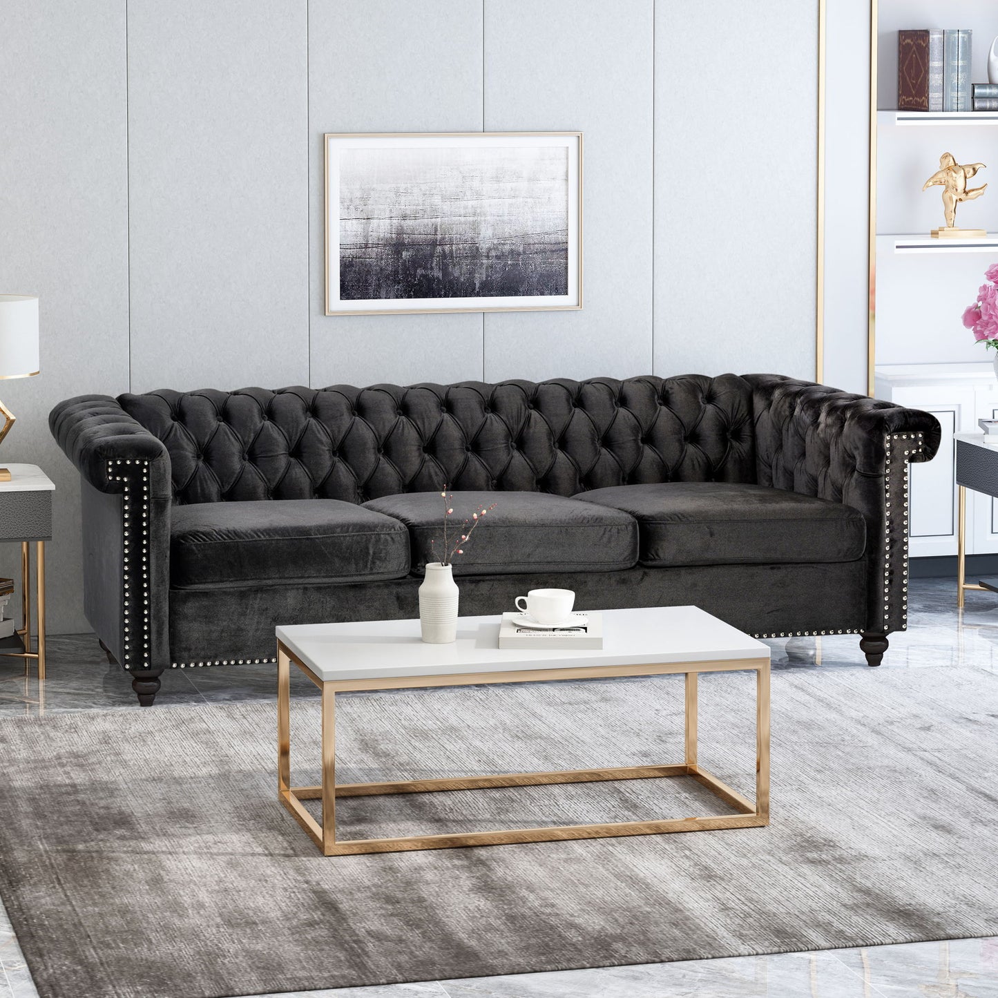 A modern living room featuring a Chesterfield Style 3 Seater Sofa With Deep Button Tufting, a white and gold coffee table, gray rug, abstract wall art above the sofa, and side shelves with decorative items and books.