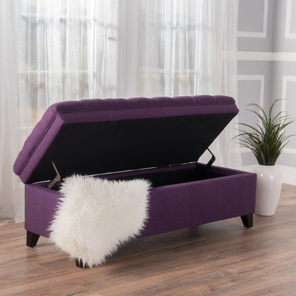 The Juliana - Storage Ottoman With Birch Legs, shown in purple upholstery with its lid open to reveal empty storage, features a white faux fur throw on one corner and sits in a bright room near sheer curtains and a potted plant.