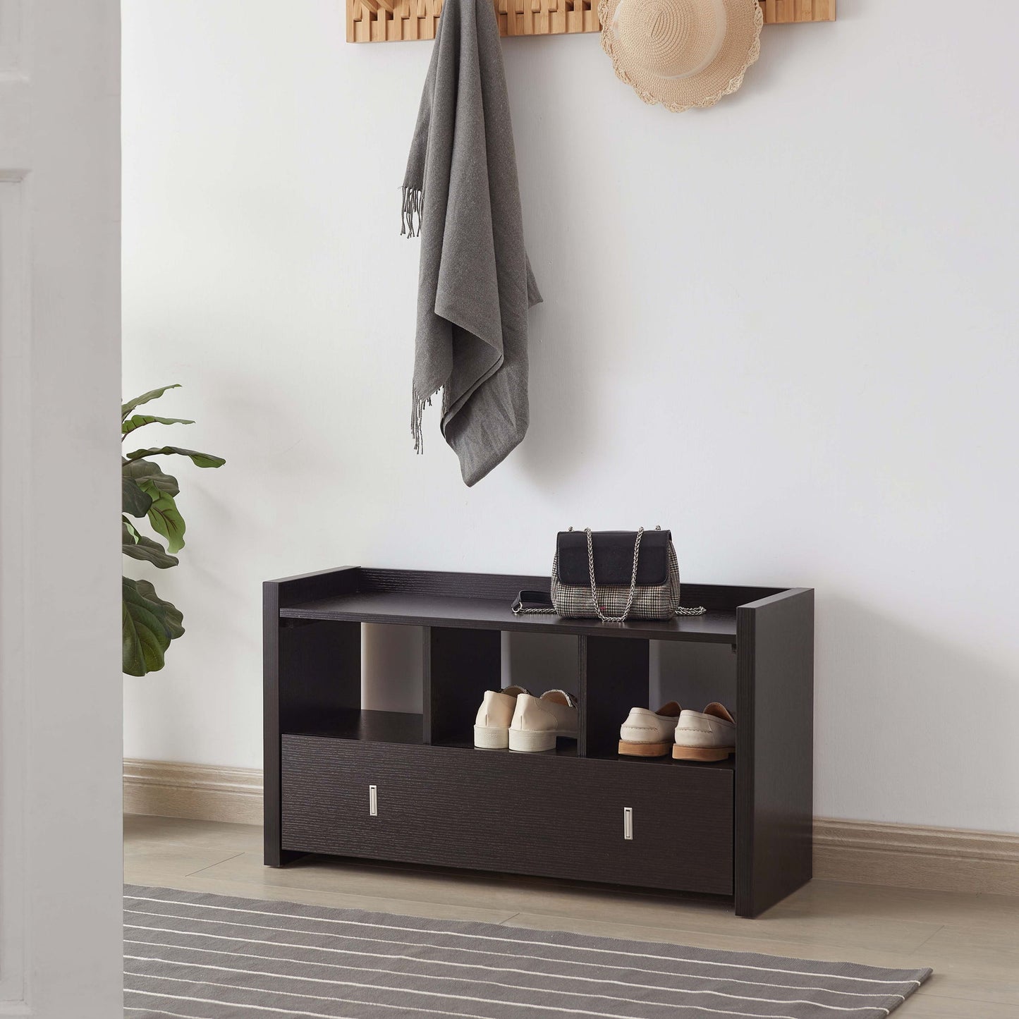A Shoe Entry Bench With Three Shelves and One Drawer With Divided Compartment holds shoes and a purse. Above it, a wooden wall rack displays a gray scarf and tan hat. A plant and striped rug are nearby.