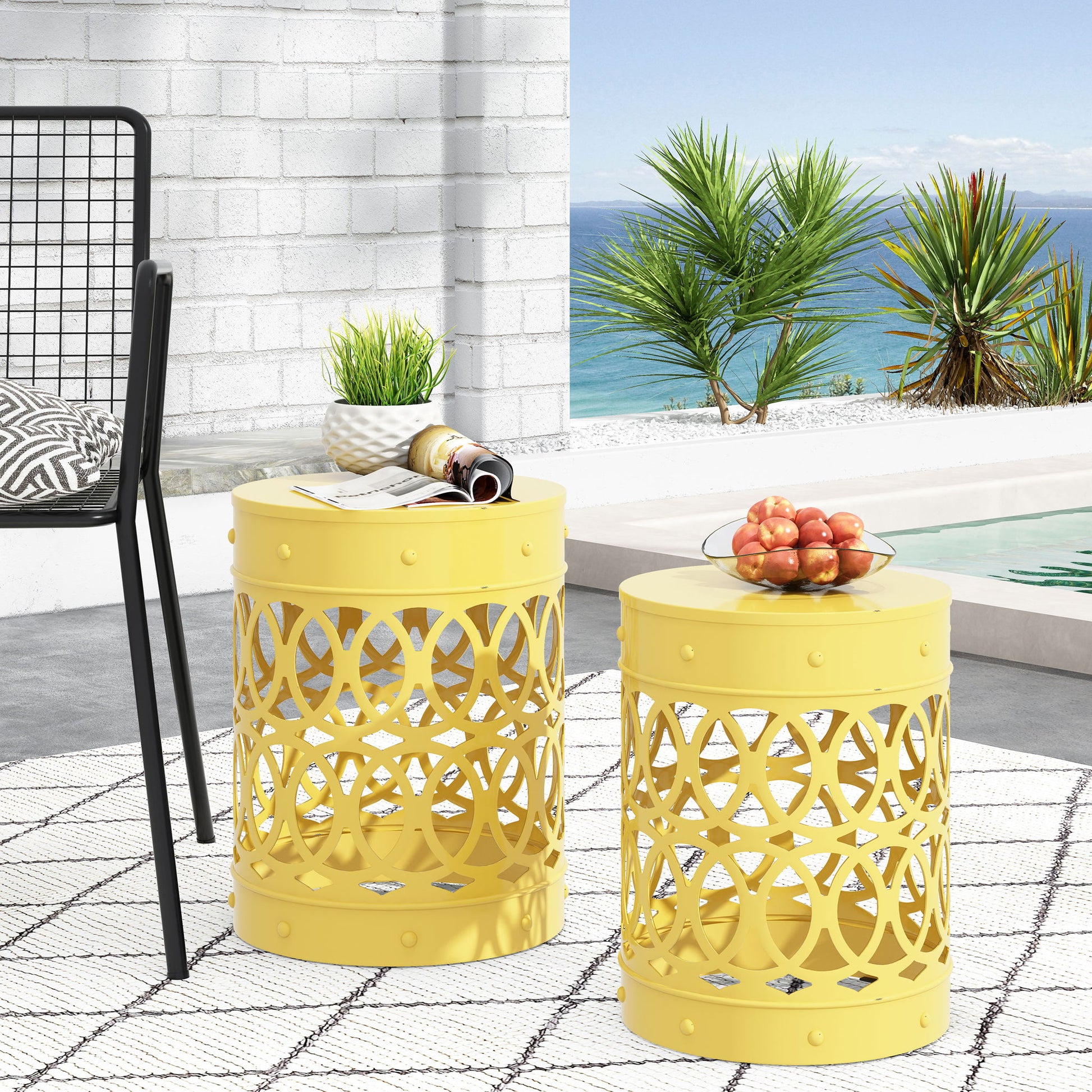 The Outdoor Metal S/2 End Table set, featuring yellow lattice designs, sits on a geometric rug by the pool with apples and magazines on top. Palm trees and the ocean backdrop create a bright coastal atmosphere.