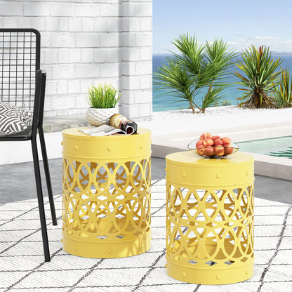 The Outdoor Metal S/2 End Table set, featuring yellow lattice designs, sits on a geometric rug by the pool with apples and magazines on top. Palm trees and the ocean backdrop create a bright coastal atmosphere.