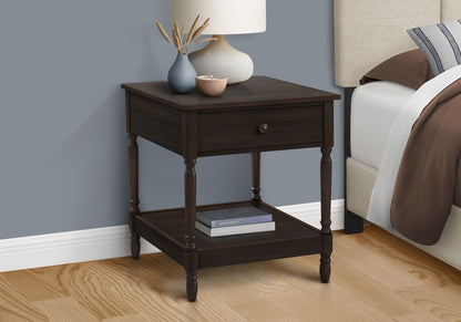 A walnut Accent 2 Tier Square Traditional table stands beside a bed with a beige headboard, topped with a white lamp, small vase of dried flowers, and bowl. A book rests on the lower shelf. The wall behind is painted gray.