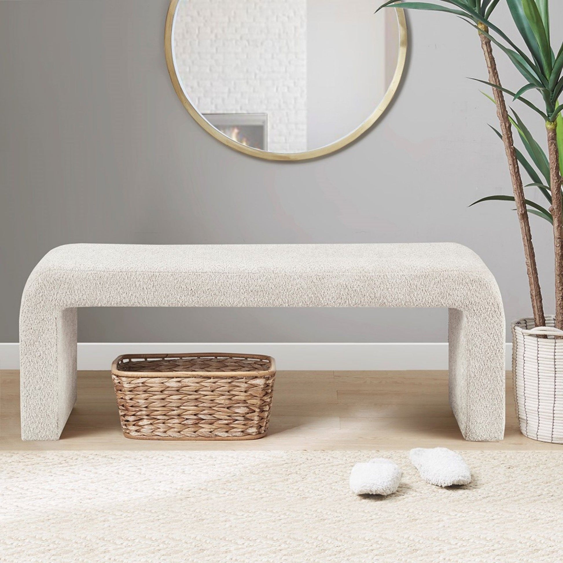 The Modern Contemporary Waterfall Bench - Cream sits on a wooden floor with a woven basket underneath. Above, a round mirror hangs on the gray wall, with a green plant and fluffy slippers nearby.